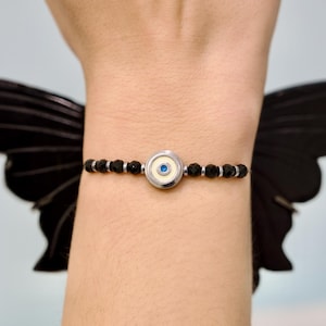 Può includere: Un braccialetto di perline nere con un ciondolo d'argento con un disegno a occhio blu. Il braccialetto è indossato su un polso, con un'ala di farfalla nera sullo sfondo. Il braccialetto è un gioiello.