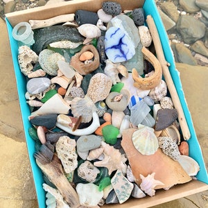 May include: A cardboard box filled with a variety of sea glass, shells, and other beach treasures. The box is lined with blue paper and the items are arranged in a random pattern.
