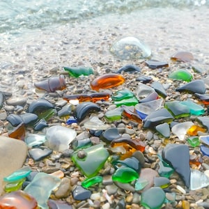 Puede incluir: Primer plano de una playa con vidrio de mar liso y colorido esparcido por la arena. El vidrio es de varios tonos de verde, azul, marrón y transparente. El agua está en el fondo.