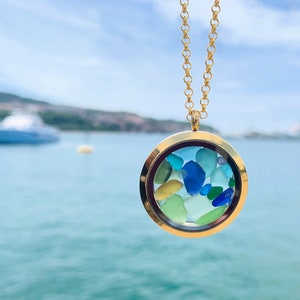 May include: Gold locket necklace with a round glass charm filled with colorful sea glass. The necklace is photographed against a blurred background of a body of water.