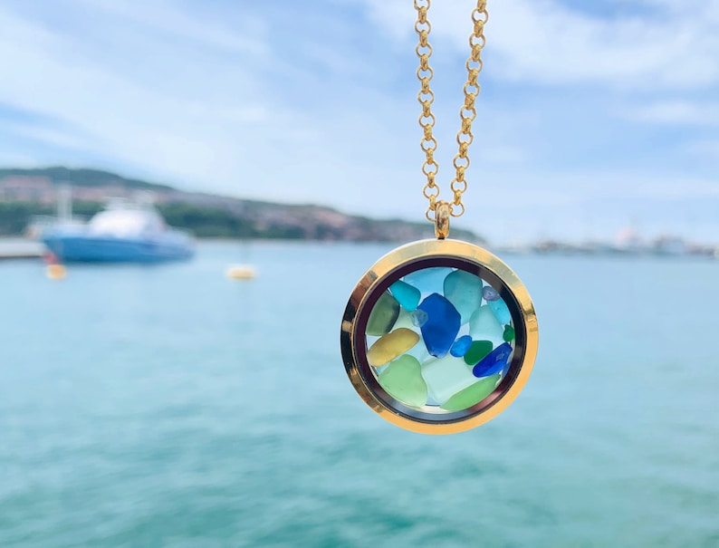 May include: Gold locket necklace with a round glass charm filled with colorful sea glass. The necklace is photographed against a blurred background of a body of water.
