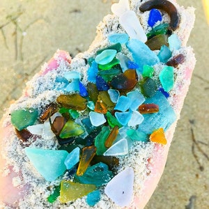 May include: A hand holding a variety of sea glass in different colors, including blue, green, brown, and white. The sea glass is mixed with sand.