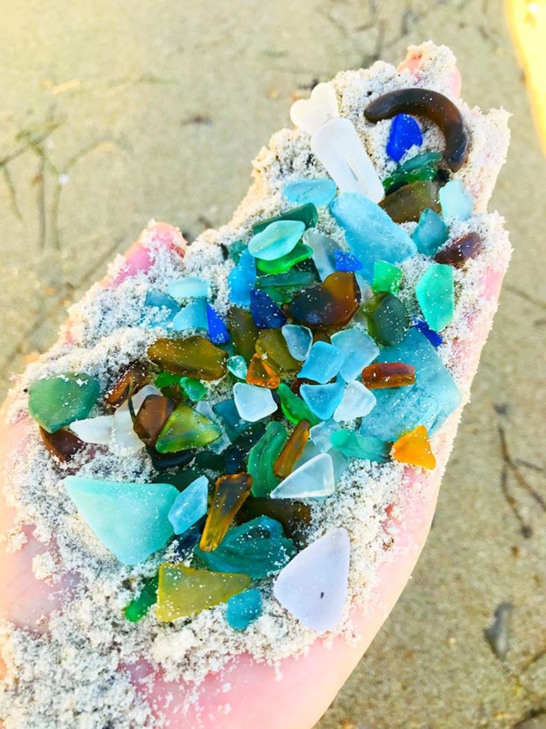 May include: A hand holding a variety of sea glass in different colors, including blue, green, brown, and white. The sea glass is mixed with sand.