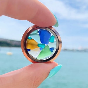 May include: A round locket with a clear glass insert filled with colorful sea glass pieces in shades of blue, green, and yellow. The locket is held in a hand against a blurred background of a body of water.