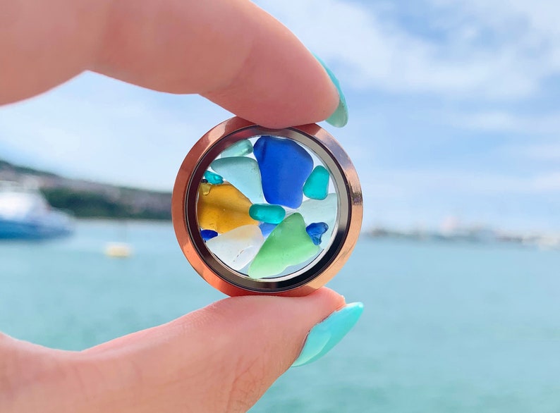 May include: A round locket with a clear glass insert filled with colorful sea glass pieces in shades of blue, green, and yellow. The locket is held in a hand against a blurred background of a body of water.