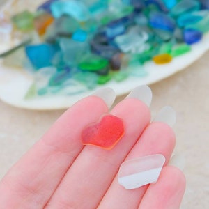 May include: Two pieces of sea glass, one is a red heart shape and the other is a clear, white, irregular shape.