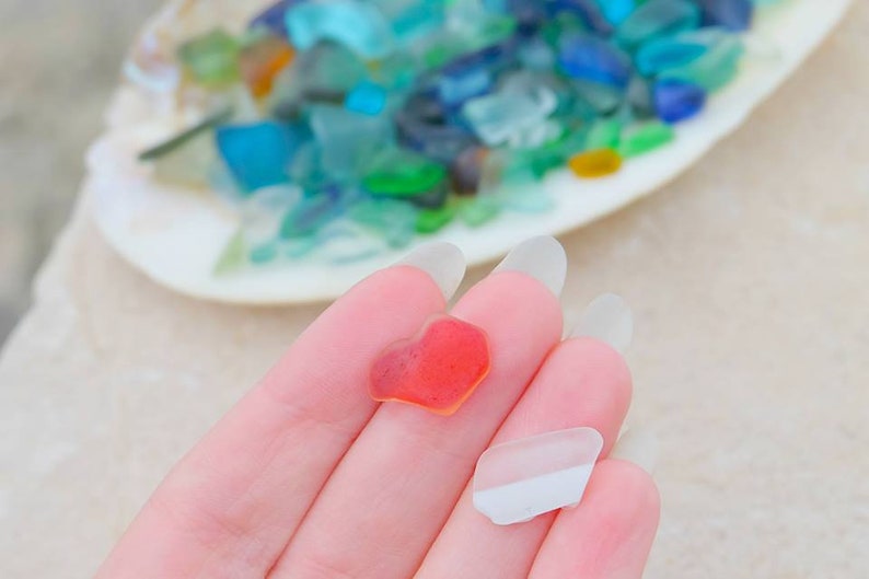 May include: Two pieces of sea glass, one is a red heart shape and the other is a clear, white, irregular shape.