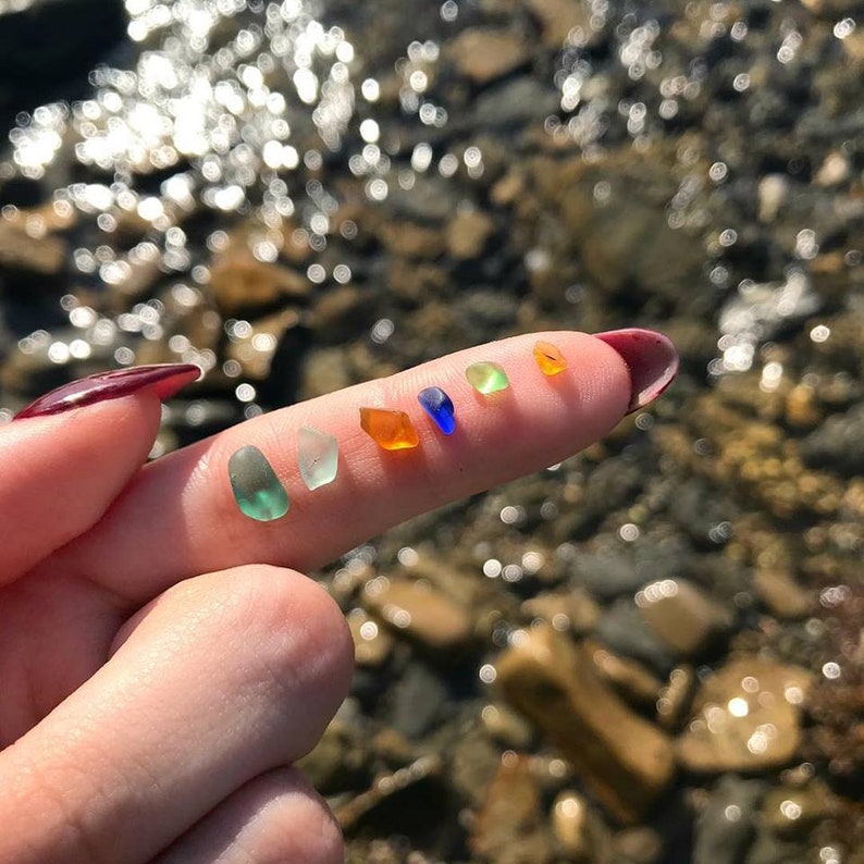 May include: A fingertip displays a collection of seven small, colorful pieces of sea glass. The sea glass is arranged in a line from left to right, starting with a light green piece, followed by a darker green piece, a light blue piece, a dark blue piece, a light orange piece, a darker orange piece, and a yellow piece.