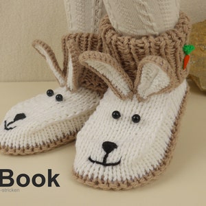 Op de afbeelding: Een paar witte gebreide konijnenpantoffels met bruine manchetten en zwarte ogen. De pantoffels hebben een wortel-applicatie op een manchet. De tekst "E-Book" en "kreatives-stricken" is zichtbaar onder de pantoffels.