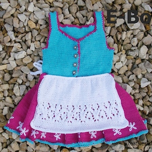 May include: A knitted baby dress with a turquoise bodice, white apron, and pink skirt. The dress has a white lace trim and a pink and turquoise trim around the neckline. The dress is laid out on a bed of small stones. The text "E-Book" is visible in the upper right corner of the image.