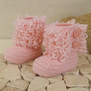 May include: A pair of pink crocheted baby booties with a fluffy, ruffled trim and two pink buttons on each bootie.