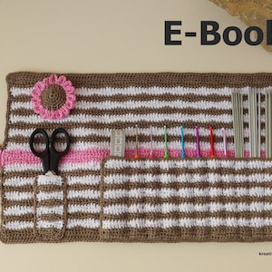 May include: A brown and white striped crochet knitting needle roll with a pink flower, scissors, and a ruler. The roll is rolled up and has a pink strap. The text "E-Book" is at the top of the image. The text "kreatives-stricken" is at the bottom of the image.