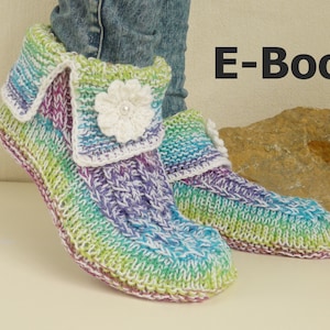 Op de afbeelding: Een paar kleurrijke gebreide pantoffels met een wit gehaakt bloemetje. De pantoffels zijn gemaakt met een regenboogkleurig garen en hebben een witte boord. De tekst "E-Book" is zichtbaar in de afbeelding.