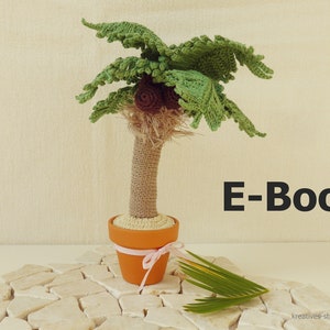 Op de afbeelding: Een gehaakte palmboom met een bruine kokosnoot en een groene pot. De pot is vastgebonden met een roze lint. De boom staat op een wit stenen oppervlak. De tekst "E-Book" is in zwarte letters op een beige achtergrond geschreven.