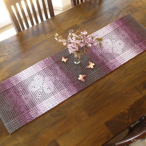 May include: A crocheted table runner in shades of gray, white, and pink with a delicate floral pattern. The runner is laid on a wooden table with a vase of pink flowers and decorative butterflies.