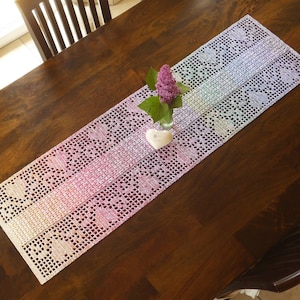 May include: A rainbow crochet table runner with a delicate floral pattern. The runner is laid on a wooden table with a vase of purple flowers in the centre.