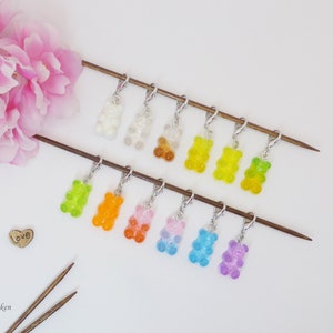 Könnte beinhalten: Ein Set mit 10 bunten Gummibärchen-Maschenmarkierern, jeder mit einem silbernen Metallring. Die Markierer sind auf zwei Holzstricknadeln angeordnet, im Hintergrund eine rosa Blume und ein Holzhäuschen mit dem Wort "love" darauf.