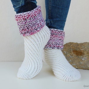 Op de afbeelding: Witte gehaakte pantoffels met roze en witte gestreepte manchetten. De sokken worden gedragen op een paar voeten met blauwe jeans.