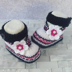 May include: A pair of white knitted baby booties with black and pink stripes, a black knitted cuff, and a white crocheted flower with a pink center and a pearl bead.