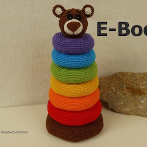 Puede incluir: Un juguete de apilar de crochet de colores arcoíris con una cabeza de oso marrón en la parte superior. El juguete está hecho de hilo y es un juguete divertido y educativo para niños. La imagen también incluye el texto "E-Book".