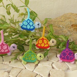 May include: A selection of crocheted turtle keyrings in various colours. Each turtle has a different coloured shell, such as blue, pink, green, yellow, and purple. The keyrings are shown against a background of foliage and stone.
