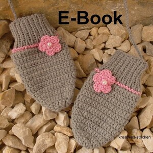 May include: A pair of gray crocheted mittens with pink flower embellishments. The mittens are lying on a bed of small gray and brown rocks. The text "E-Book" is visible above the mittens.