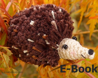 May include: A brown and white crocheted hedgehog with black eyes and a black nose. The hedgehog is sitting on a branch of a tree with orange leaves. The text "E-Book" is written in black letters on a white background.