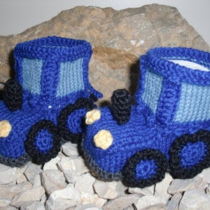 Puede incluir: Un par de botines de bebé de punto azul con forma de tractor. Los botines tienen ruedas negras, faros amarillos y una cabina de punto azul y gris.