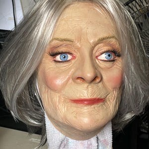 Könnte beinhalten: Eine realistische Maske einer älteren Person mit heller Haut, blauen Augen und grauem Haar. Die Maske hat detaillierte Falten, rosige Wangen und roten Lippenstift. Sie liegt auf einem weißen Handtuch.