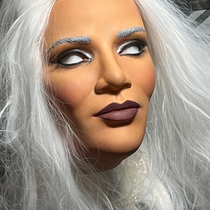 Op de afbeelding: Een volledig masker met een gebruinde teint, lang wit haar en zilveren wenkbrauwen. De ogen zijn effen wit met zwarte eyeliner en de lippen zijn geschilderd in een matte donkerpruime kleur. Het masker heeft een gladde, gebeeldhouwde gezichtsvorm.