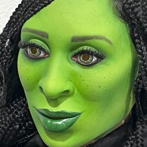 Puede incluir: Primer plano de un rostro con piel verde, labios verdes y cabello oscuro trenzado. Los ojos son de color claro con pestañas oscuras. La persona lleva una prenda negra.