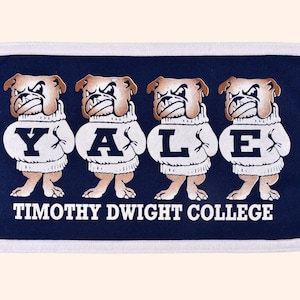 May include: A navy blue and white rectangular towel featuring four bulldog illustrations wearing sweaters that spell out "YALE". The text "TIMOTHY DWIGHT COLLEGE" is printed below the bulldogs.