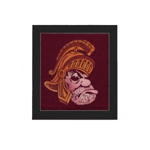 May include: Framed artwork featuring a stylized illustration of a Roman soldier's head in a golden helmet. The artwork has a burgundy background and is set within a black frame. The soldier's face is detailed with a stern expression.