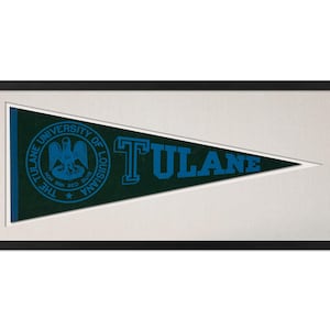 May include: Framed Tulane University pennant. The pennant is a dark green triangle with the word "TULANE" in blue block letters. The university seal is on the left side. The pennant is framed in a black frame.