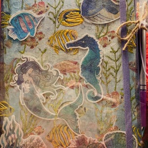 May include: A journal with a mermaid and sea life theme. The cover features a mermaid, seahorse, whale, and various fish stickers. The background has an underwater scene with seaweed and glitter accents. A purple ribbon and twine are attached.