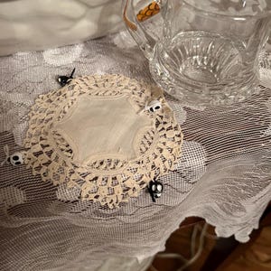Puede incluir: Una jarra de crema de vidrio transparente con un diseño naranja y negro en el asa se encuentra junto a un tapete de encaje color crema decorado con pequeños alfileres de calavera. El tapete descansa sobre un mantel de encaje blanco.