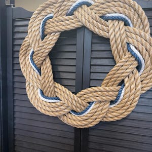 May include: A circular wreath constructed from thick, tan-colored natural fiber rope. The rope is woven into a continuous knot pattern, accented with thin, intertwined strands of navy blue and white cord that follow the circular design.