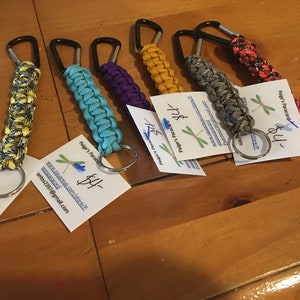 May include: A collection of colorful paracord keychains with metal clips and key rings. The keychains are made with different colored paracord and have a variety of patterns. The keychains are priced at $4 each. The text on the cards reads "Paige's Paracord" and includes a website address and email address.