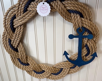 Puede incluir: Una corona de temática náutica hecha de gruesa cuerda trenzada en color beige natural y azul marino. Un ancla azul marino está unida a la corona. La corona está colgada en una pared blanca con paneles.