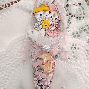 May include: A pink and white Easter cone with a floral pattern. Two white bunnies with yellow and pink accents are peeking out of the top of the cone. A banner at the bottom reads "Happy Easter".