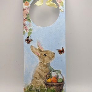 May include: Easter-themed door hanger with a light blue backdrop. It depicts a rabbit with a basket of eggs, floral details, and butterflies. A yellow ribbon and heart-shaped embellishments complete the design. Measurements in cm.