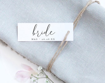Könnte beinhalten: Weiße Karte mit dem Wort "bride" in schwarzer Kursivschrift. Die Karte enthält auch den Text "M&S - 06.08.XX". Die Karte ist mit Schnur gebunden und auf einer hellblauen Serviette platziert.