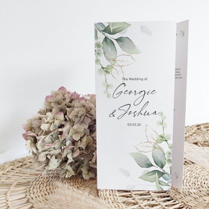 Puede incluir: Una invitación de boda doblada con un diseño de acuarela de hojas verdes y detalles dorados. El texto dice "The Wedding of Georgie & Joshua 09.03.26".