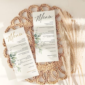 May include: Two elegant wedding menu cards with the word "Menu" in a stylish script. The cards feature a floral design with green leaves and a list of menu items. The menus are on a woven placemat with dried ornamental grass.