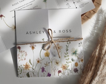 May include: A wedding invitation with a floral design. The invitation is folded in half and tied with twine. The front of the invitation has the names Ashley and Ross written in black text. The back of the invitation has a timeline of the wedding day.