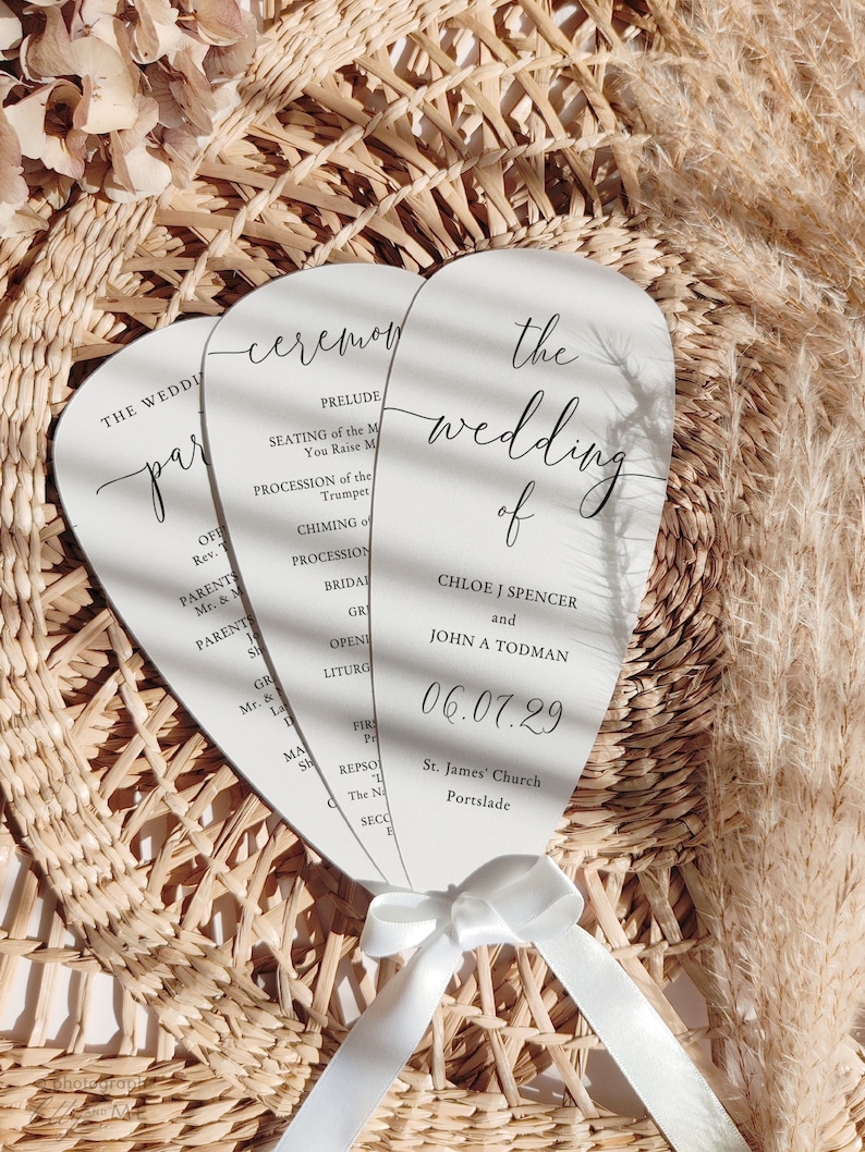May include: Three white wedding ceremony programs with a curved top, tied with a white ribbon. The programs have black text, including the wedding date, names, and ceremony details. The background is a woven basket.