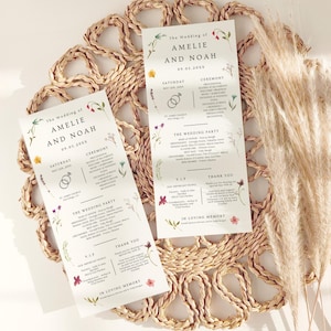 Puede incluir: Dos programas de boda con detalles florales y los nombres "AMELIE Y NOAH" impresos. Los programas incluyen detalles de la ceremonia, nombres del cortejo nupcial y un mensaje de agradecimiento. Sobre un salvamanteles tejido.