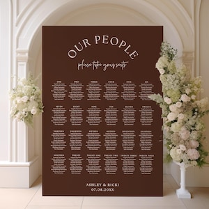 May include: A dark brown wedding seating plan with white lettering. The words "OUR PEOPLE" and "Please take your seats" are at the top. Names are listed in numbered columns. The names Ashley & Ricki and the date are at the bottom.