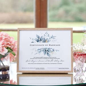 May include: A framed certificate of marriage with a watercolor floral design in shades of blue and gold. The text reads "Certificate of Marriage" and includes the names Axelse Guzanne Harrison and Jonathan Rodacy Dixon, the date August 31, 2028, and the location St. Peter's Hall, San Francisco, California.