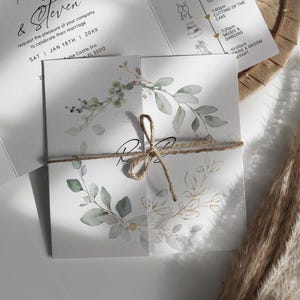 May include: Wedding invitation suite featuring a square card tied with twine. The card has watercolor botanical illustrations in green and gold. The invitation includes the names "& Steven", date, time, and location details.